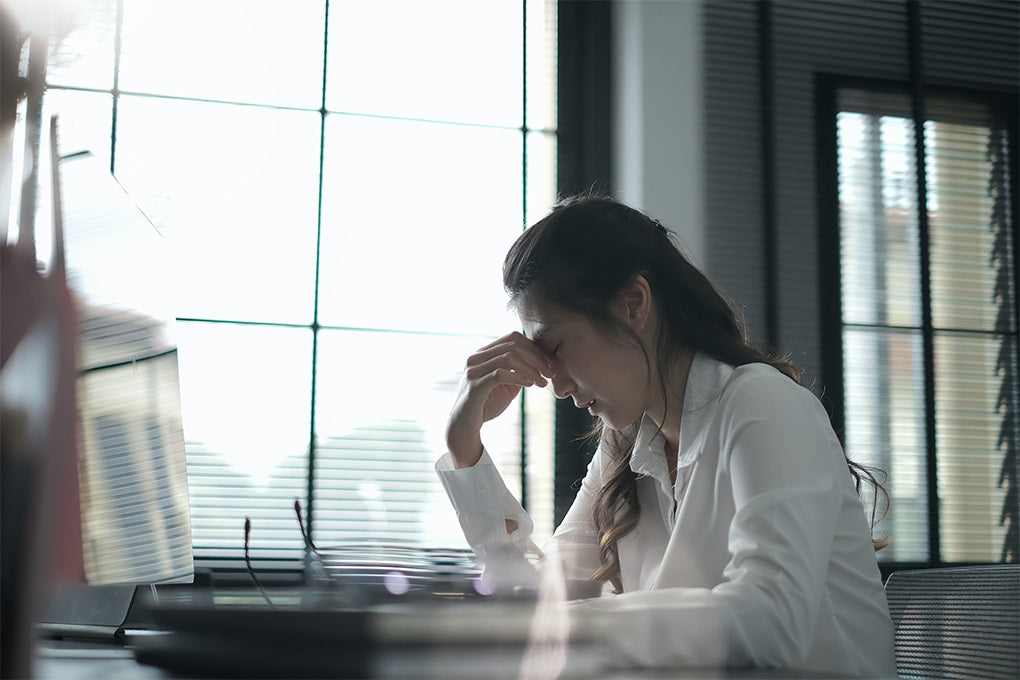 A depressed woman sitting on a chair in front of a desk with her eyglasses open and her right hand on her forehead 