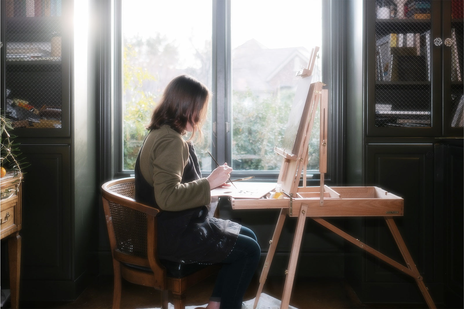 A woman sitting on a chair besides a window with drawing brushes in her hands painting on a canvas