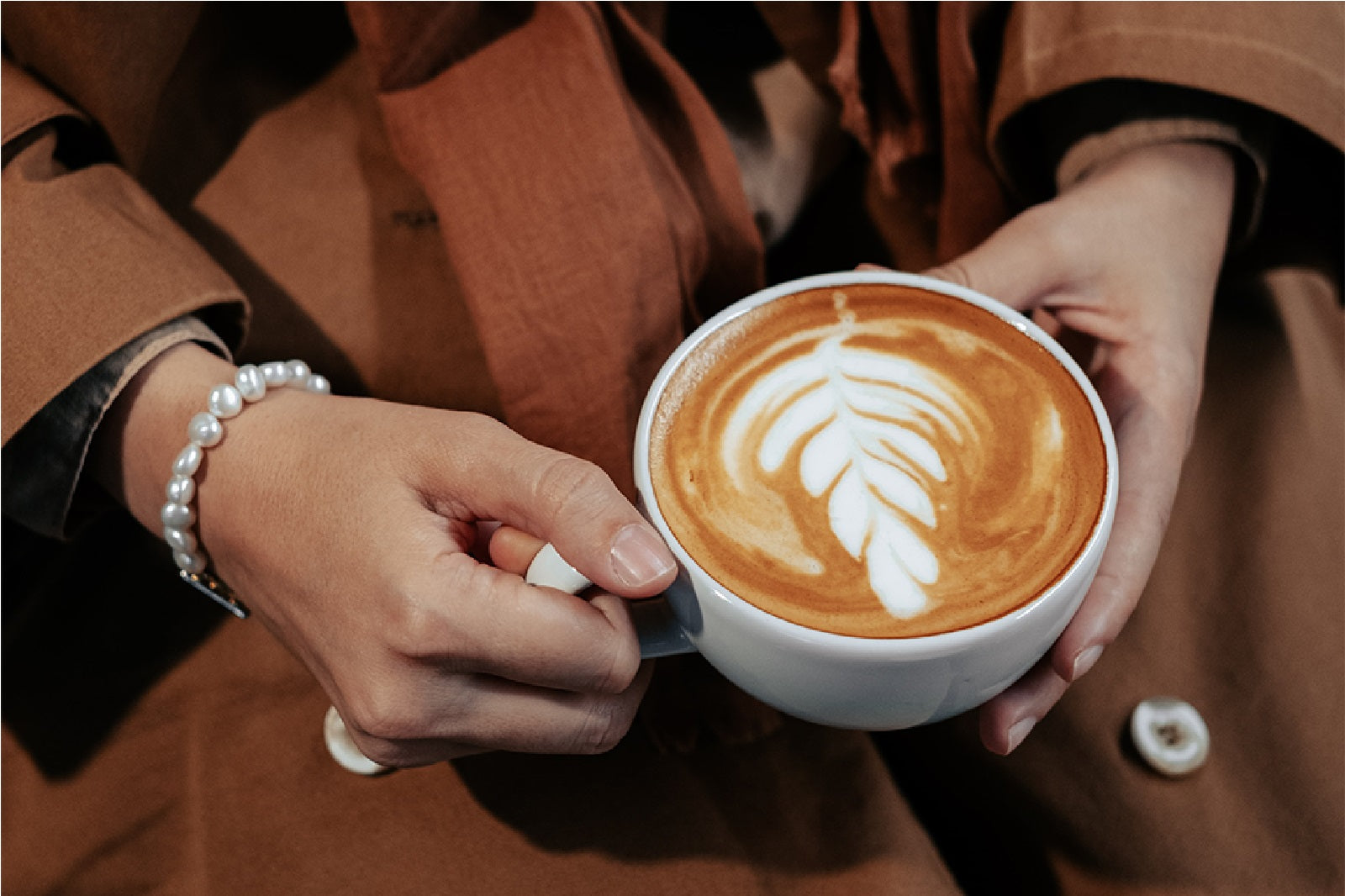A woman wearing a brown coat and a white pearl bracelet on her right hand holding a cup of cappucino with her two hands