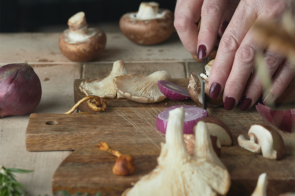A pair of hands of a wpman cutting onions and mushrooms on a wooden cutting board on a table